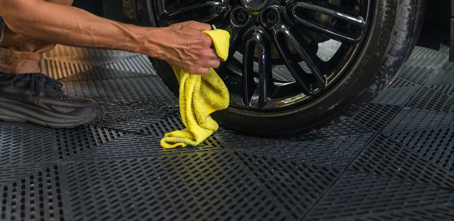 Person cleaning a car wheel with a yellow cloth on a textured black mat.