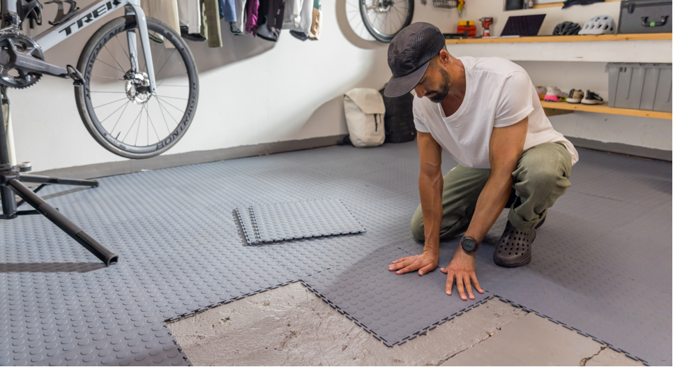 Man installing interlocking tile floor tiles in a workshop with bicycles and tools in the background.